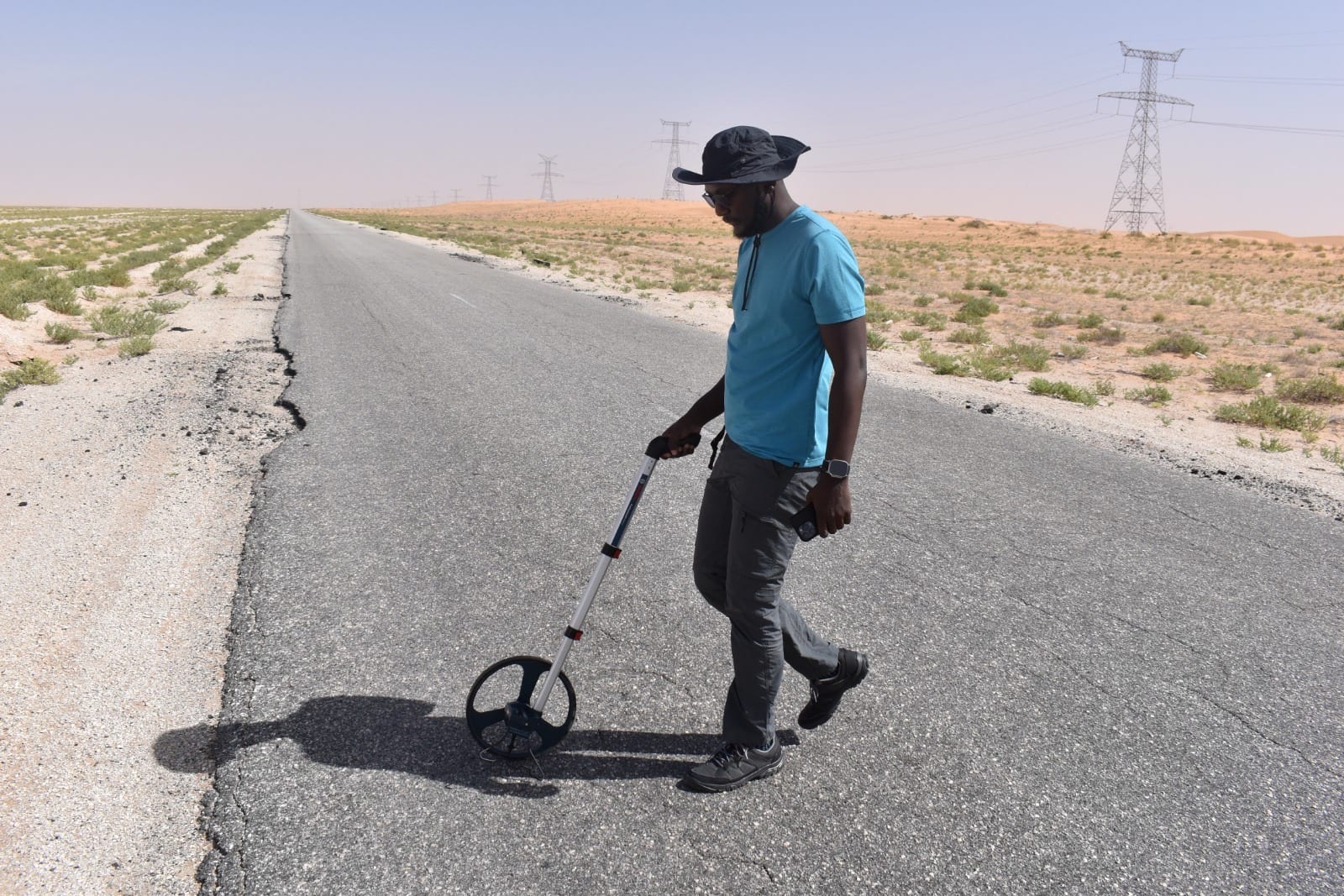 A carriageway-width survey walked with a measuring wheel along the cracked running surface of the Nouadhibou to Zouerat road, transmission towers lining the corridor