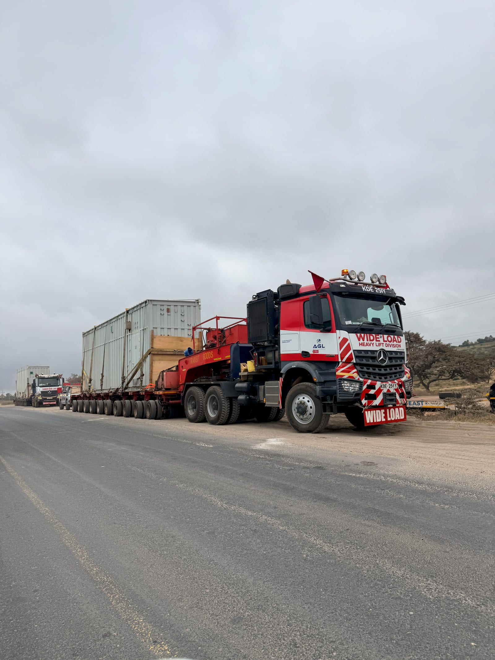 A wide-load escort tractor unit carrying a containerised module on a multi-axle modular trailer, held on the shoulder of a Kenyan highway
