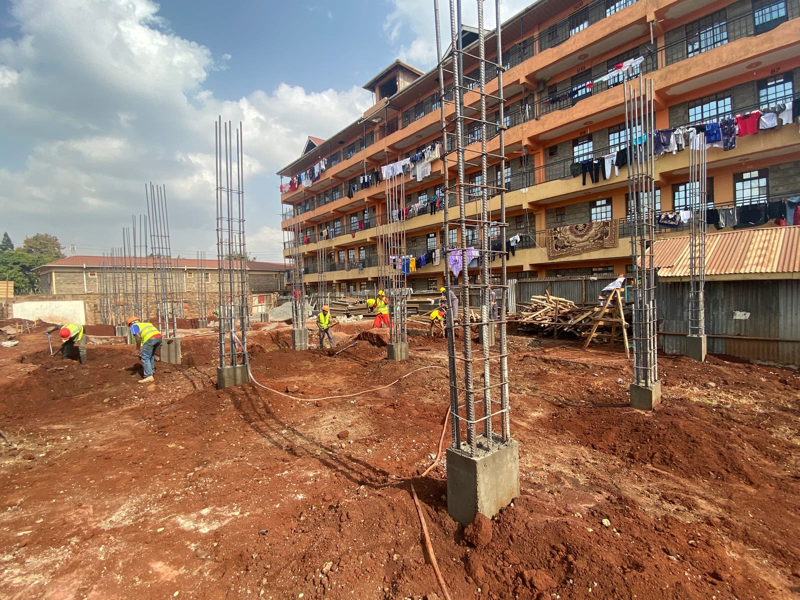 Reinforced-concrete column starter cages rising from the footings at the ACK Trinity Muthua site in Uthiru, a construction crew alongside an existing block