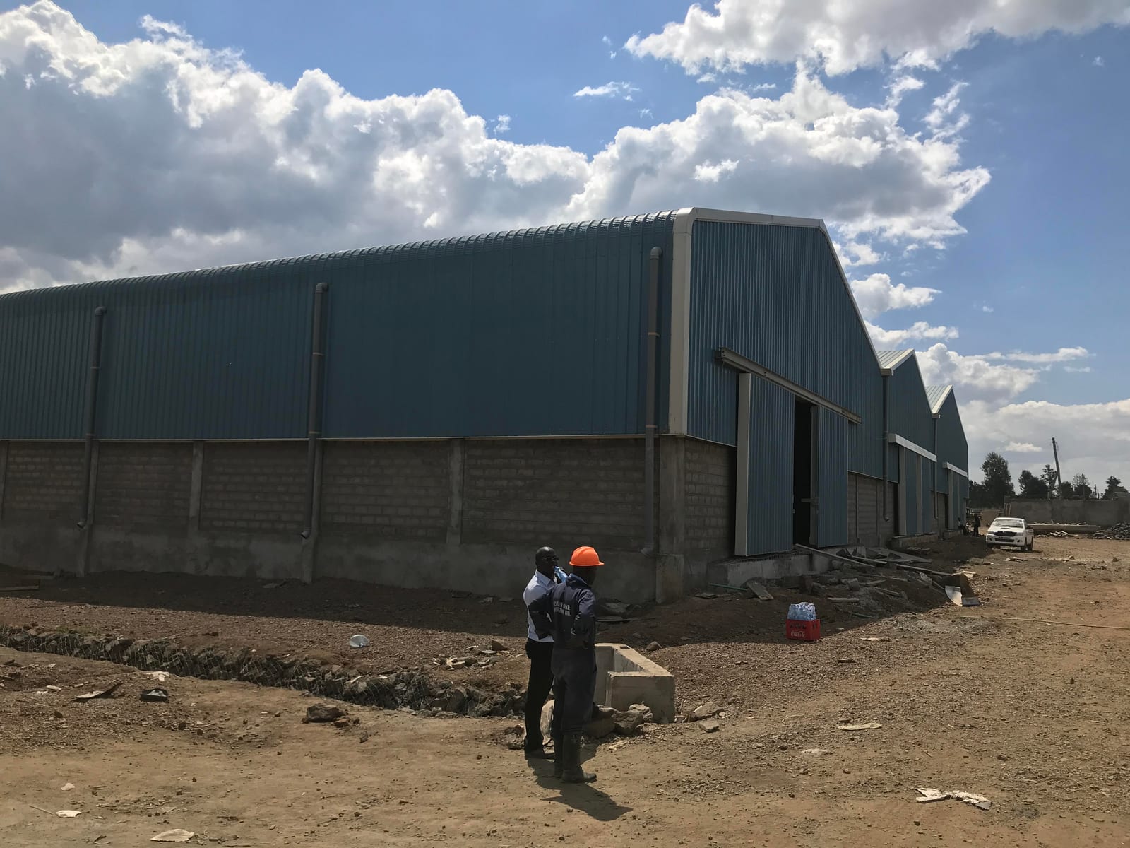 A blue-clad steel-frame warehouse under construction at VisionOne, Nakuru, two workers giving scale